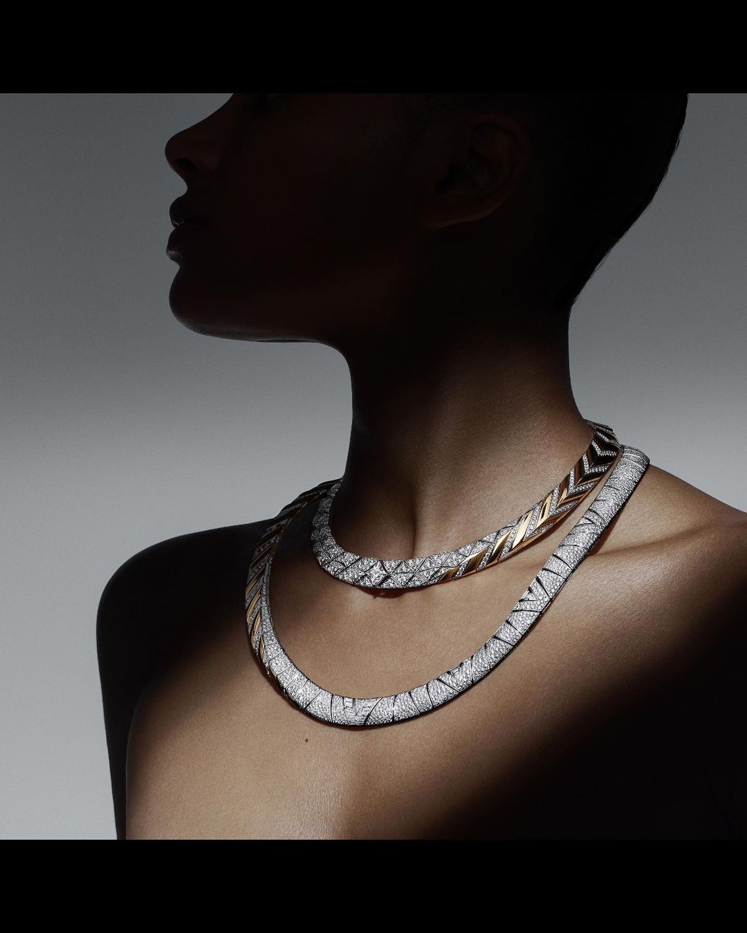 Elegant fashion shot of a woman wearing two diamond necklaces against a grey gradient background with dramatic side lighting creating shadows.