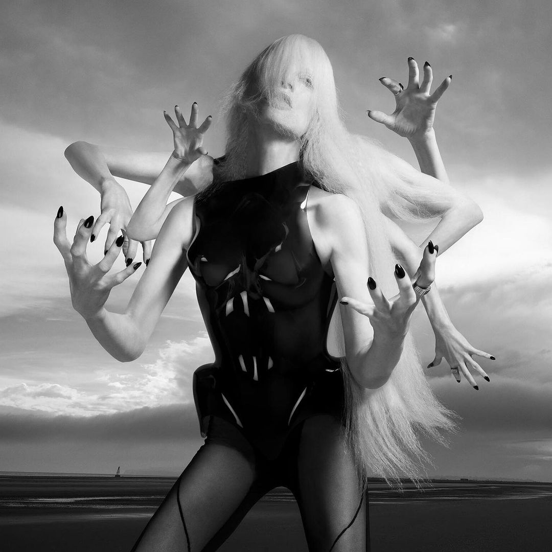 Monochrome fashion shot of a woman in a black bodysuit with multiple arms extended, black fingernails, and a long white wig against a cloudy sky.