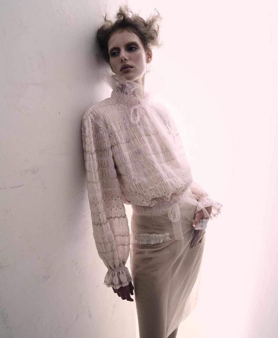 A woman poses in a sheer pink blouse and skirt with ruffles, leaning against a bright white wall in a studio-lit fashion shot.