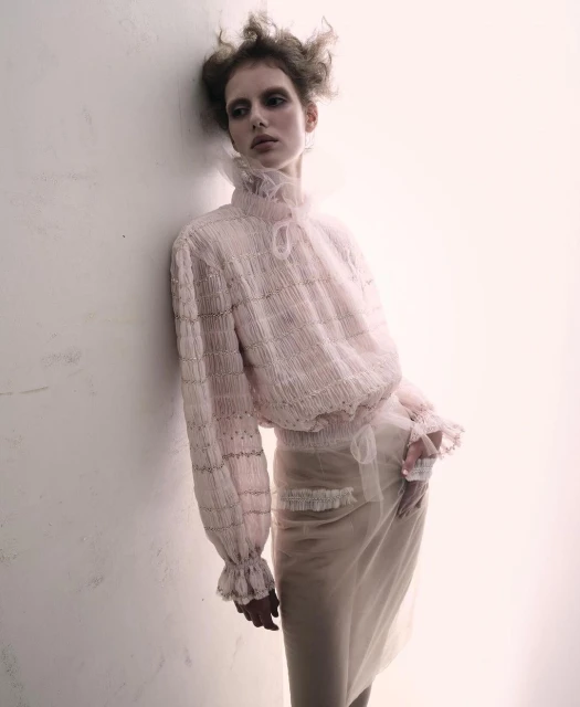 A woman poses in a sheer pink blouse and skirt with ruffles, leaning against a bright white wall in a studio-lit fashion shot.