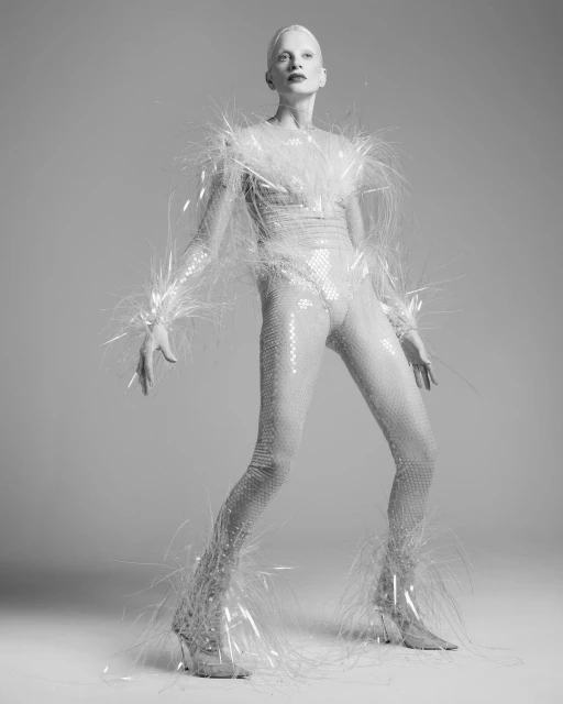 Monochrome fashion shot of a person in a sequined catsuit with feather-like trim, striking a pose against a pale grey backdrop.