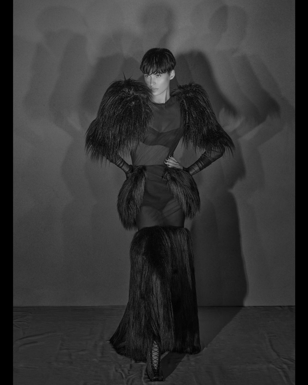 A woman poses in a dark, feathered fashion ensemble against a gray backdrop, creating a dramatic and high-contrast black-and-white image.