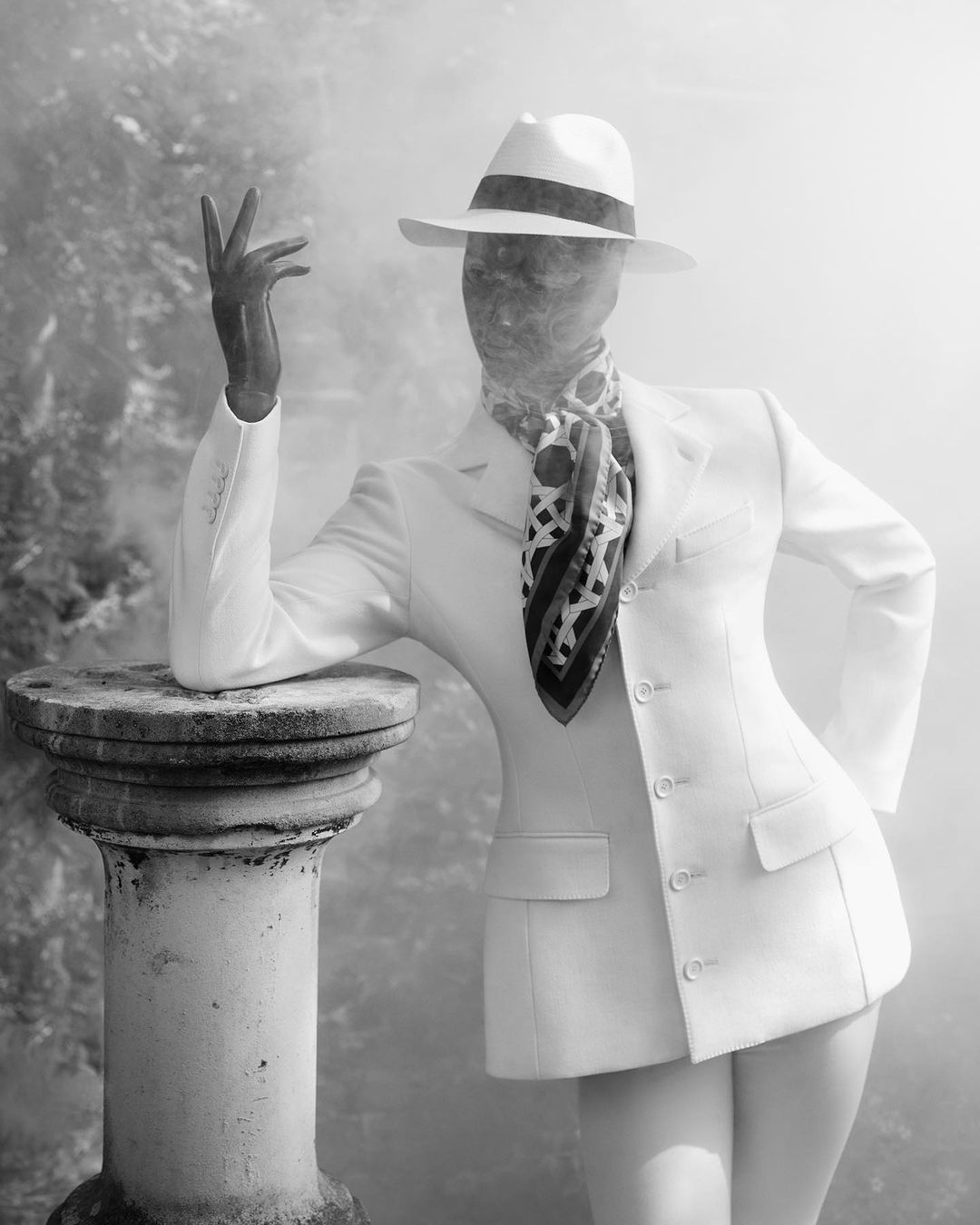 Monochrome fashion portrait of a model wearing a white suit, fedora, patterned silk scarf, and veiled face, leaning on a stone column