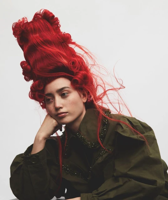 A young adult female in a green embellished jacket and red elaborate curly wig poses against a neutral background in a studio setting.