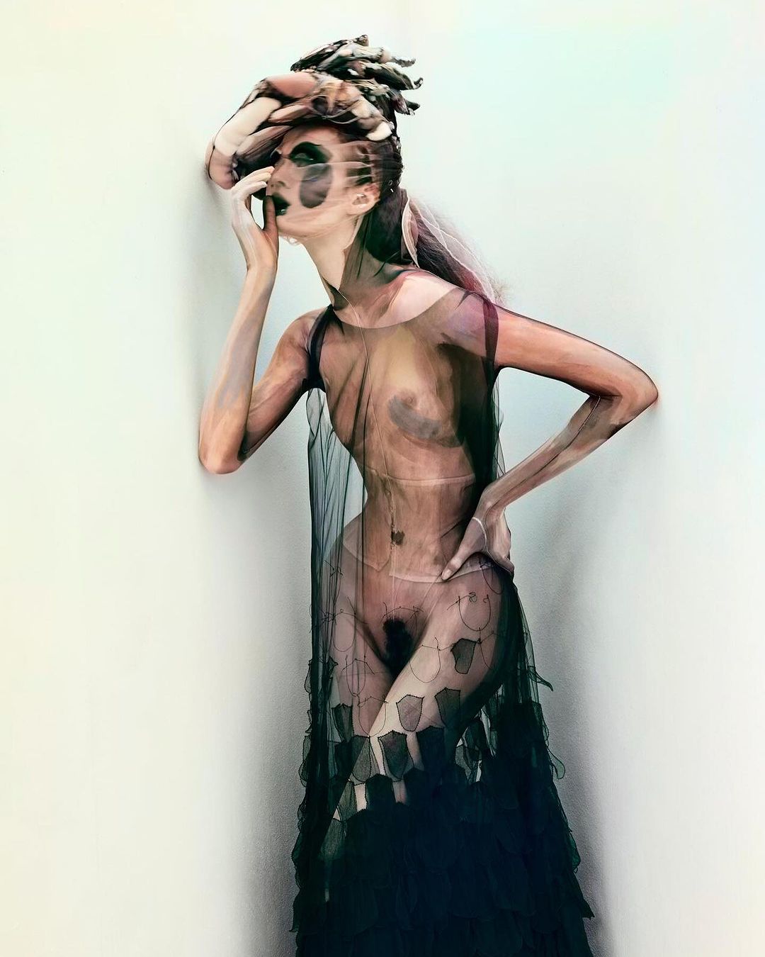 Artistic fashion portrait of a model in sheer black fabric, face makeup, and a sculptural headpiece, posing against a neutral wall backdrop.