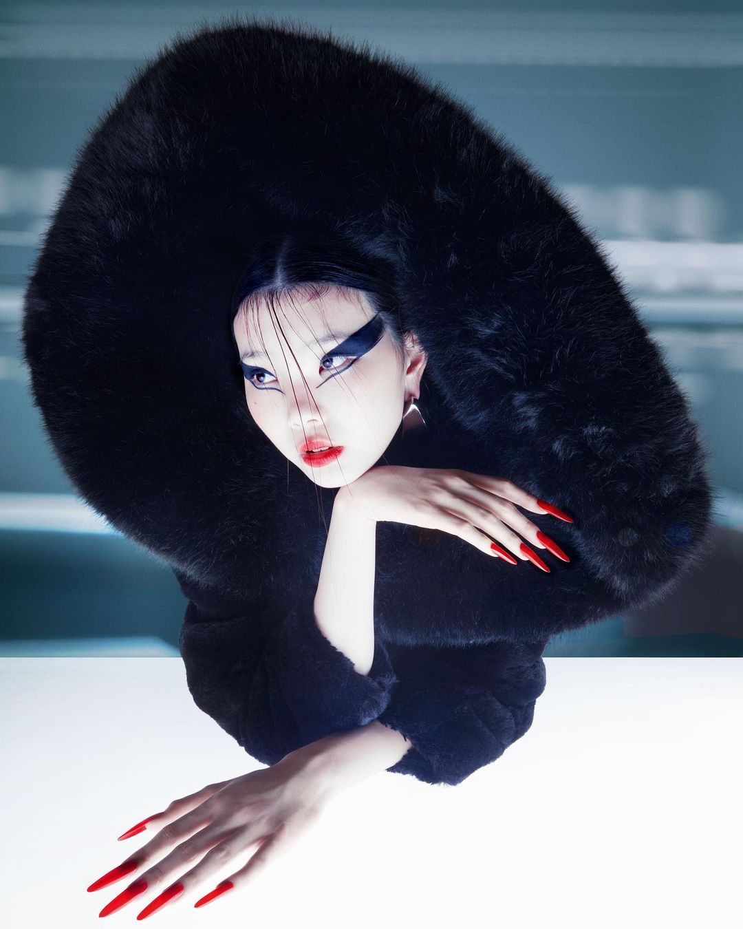 Asian woman in a black fur hooded coat with exaggerated red nails and bold makeup poses in a studio with a neutral background.