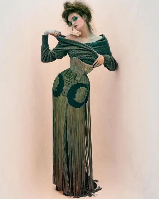 A woman wearing a vintage-style outfit with a gray sweater, a wide gold belt, and fringed pants poses in front of a peach background.