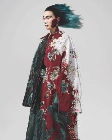 Model with vibrant teal hair and facial paint wearing a floral patterned jacket against a plain light grey studio background.