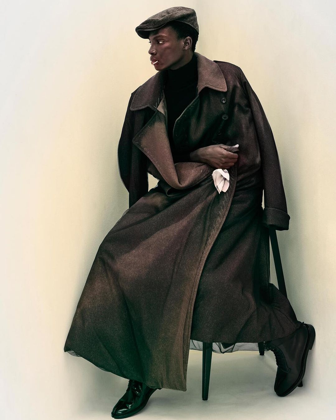 Black man wearing a brown wool coat, black turtleneck, and flat cap seated against a plain white studio background.