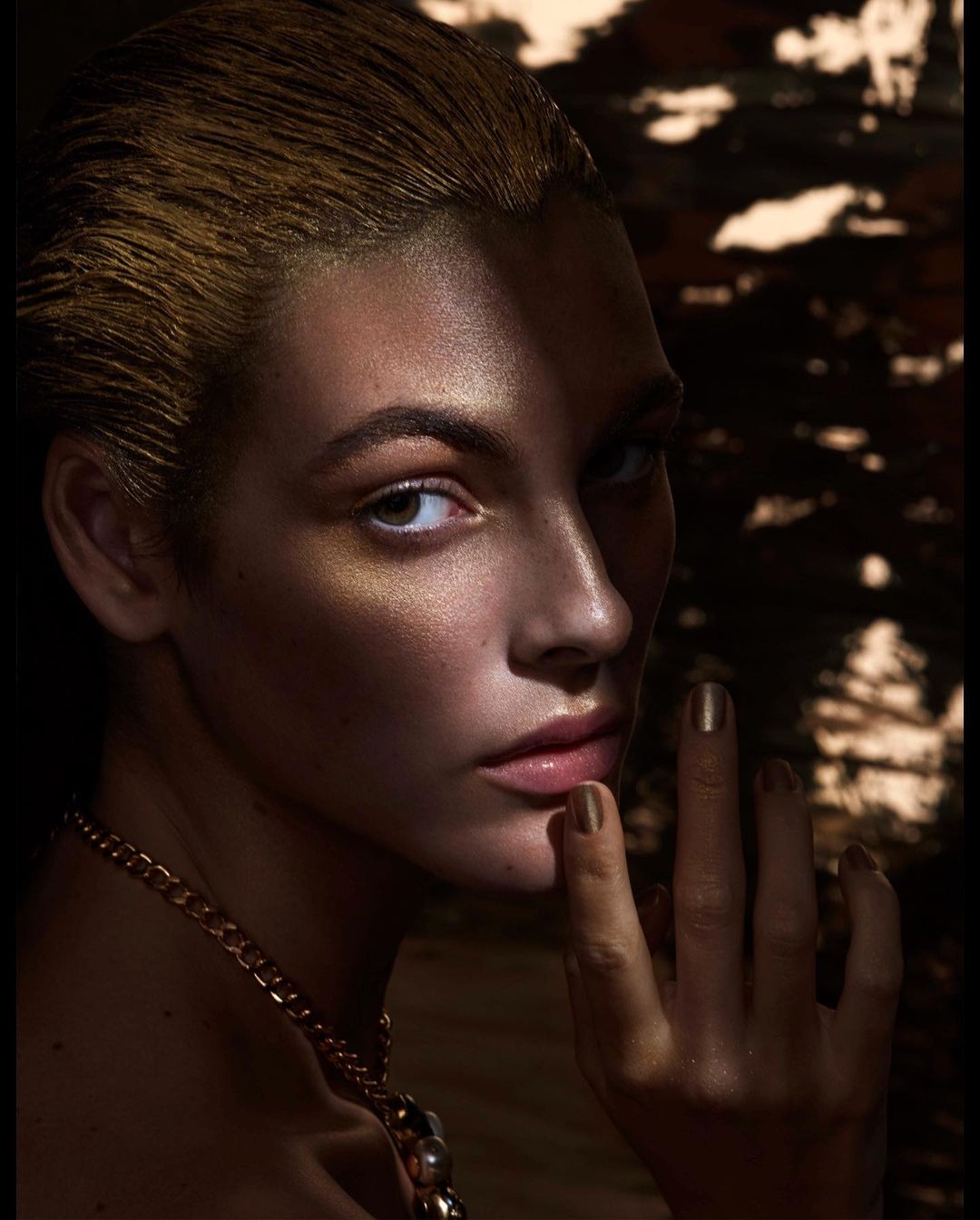 Close-up portrait of a woman with metallic gold hair, shimmering skin highlights, and a gold chain necklace by photographer Solve Sundsbo.