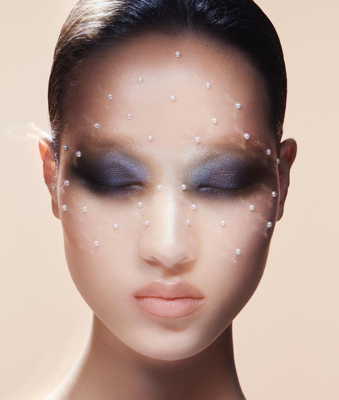 Close-up portrait of an adult female with eyes closed, adorned with small pearls on her face, set against a neutral gradient background.