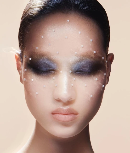Close-up portrait of an adult female with eyes closed, adorned with small pearls on her face, set against a neutral gradient background.