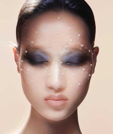 Close-up portrait of an adult female with eyes closed, adorned with small pearls on her face, set against a neutral gradient background.