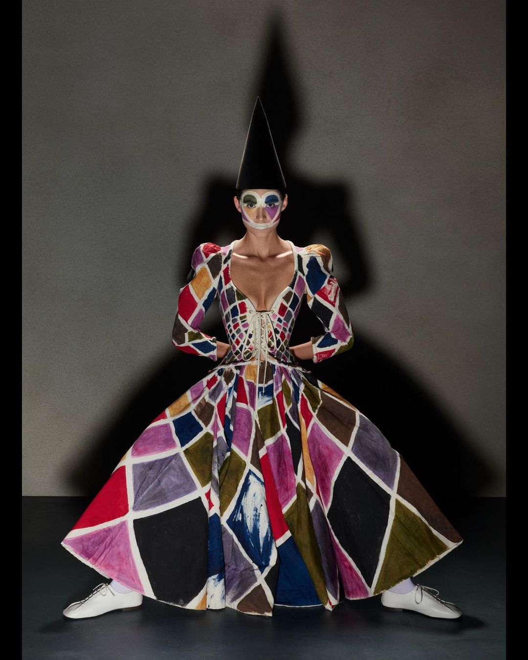 Woman in harlequin dress with face makeup and cone hat stands in studio with shadow. Fashion, art and costume design.