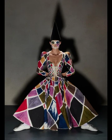 Woman in harlequin dress with face makeup and cone hat stands in studio with shadow. Fashion, art and costume design.