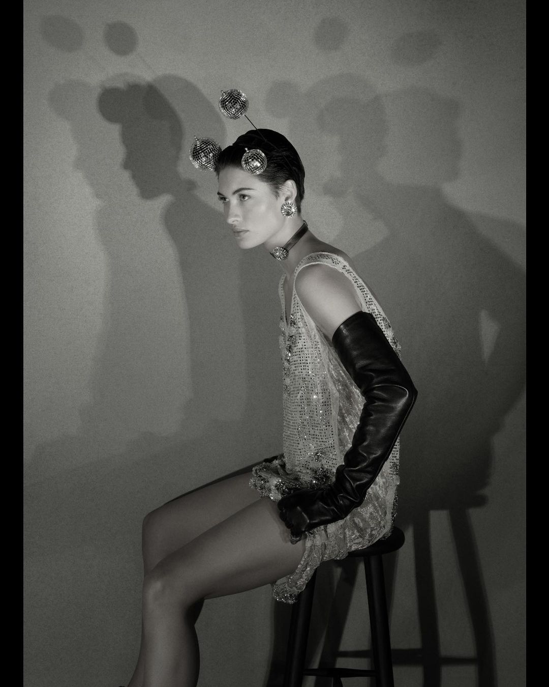 Monochrome portrait of a woman seated on a stool, wearing a beaded dress, opera gloves, and disco ball headpiece against a plain backdrop.
