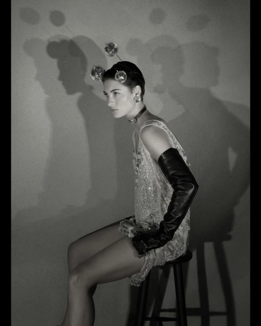 Monochrome portrait of a woman seated on a stool, wearing a beaded dress, opera gloves, and disco ball headpiece against a plain backdrop.