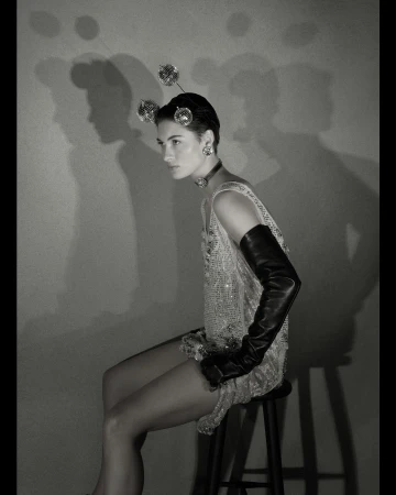 Monochrome portrait of a woman seated on a stool, wearing a beaded dress, opera gloves, and disco ball headpiece against a plain backdrop.