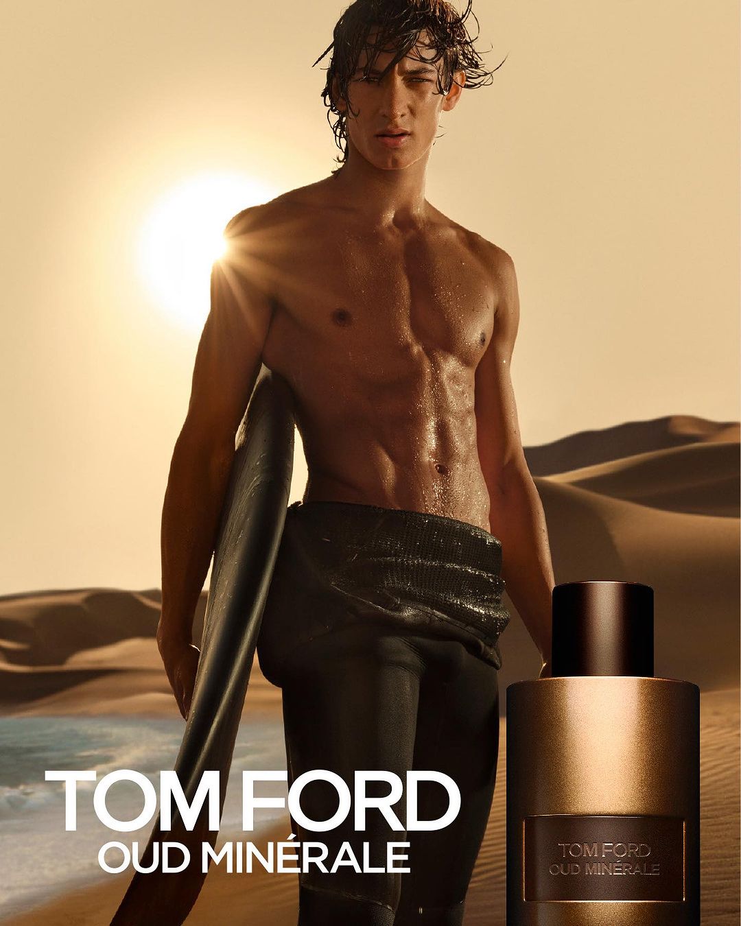 Man with surfboard poses with Tom Ford Oud Minérale perfume bottle against desert backdrop in a beauty and fashion product shot.