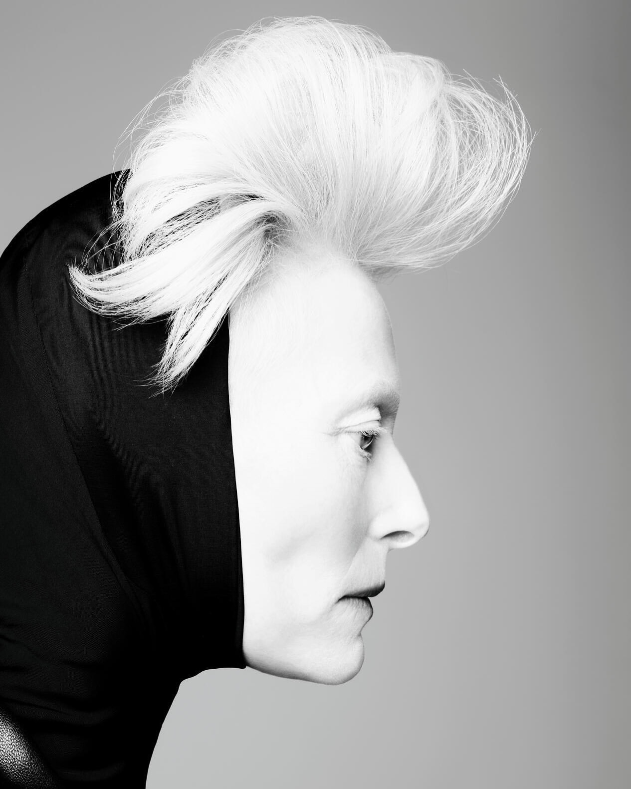 Profile portrait of a fair-skinned person with white hair styled upwards, wearing a black hood and dark lipstick, against a grey backdrop.