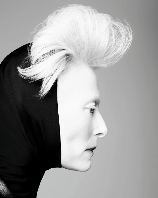 Profile portrait of a fair-skinned person with white hair styled upwards, wearing a black hood and dark lipstick, against a grey backdrop.