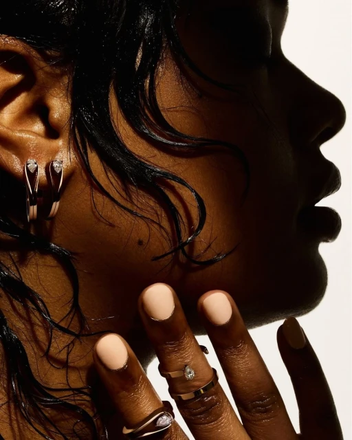 Close-up shot of a dark-skinned woman showcasing jewelry: hoop earrings and rings, with glossy dark hair and tan nail polish.