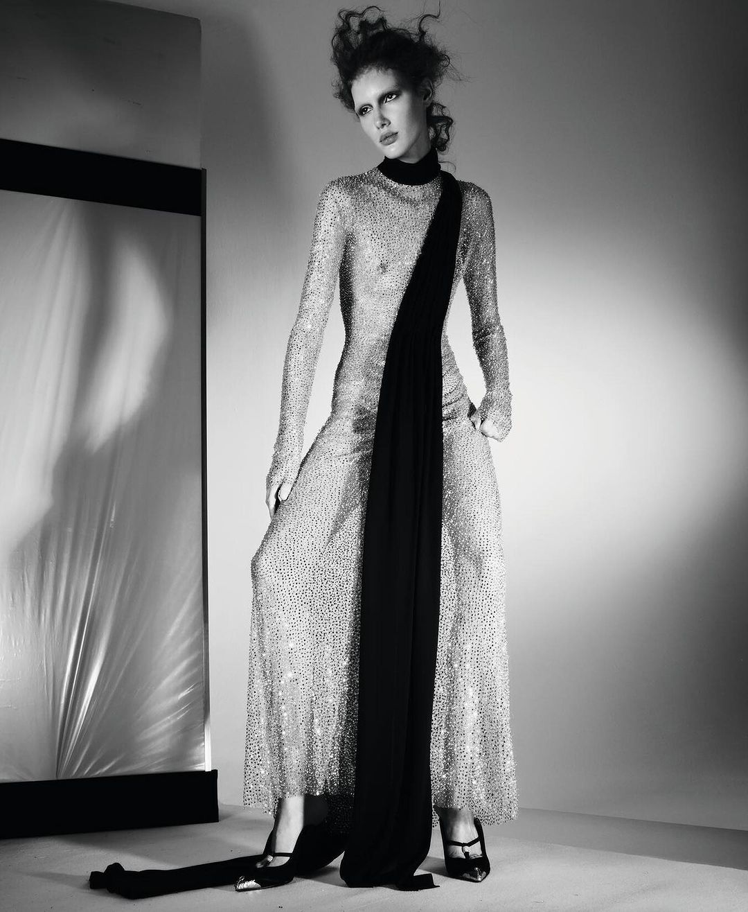 A fashion shot in black and white features a slender woman in a sequined gown with a black scarf, posing in front of a reflective panel.