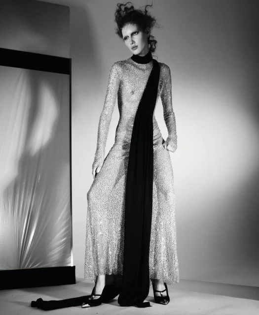 A fashion shot in black and white features a slender woman in a sequined gown with a black scarf, posing in front of a reflective panel.