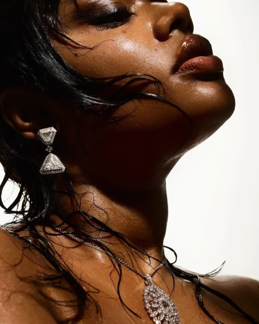Close-up of a Black woman with glossy skin, wet hair, and triangular diamond earrings looking up against a stark white background.