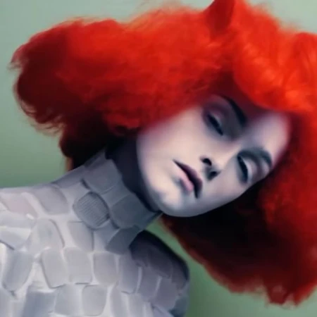 Woman with bright red curly hair and pale skin poses in structured white top against a light green background in an artistic portrait.