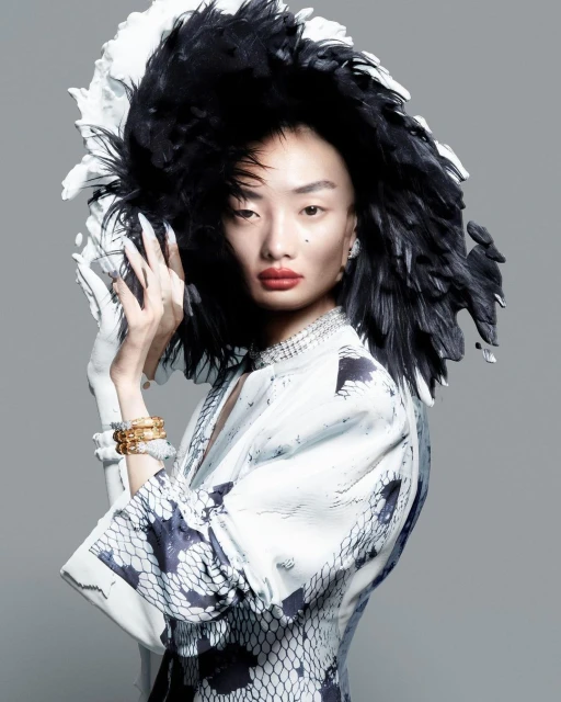 Portrait of an Asian woman with dramatic makeup and a large black-and-white feathered headpiece, posing in a light-colored floral dress.