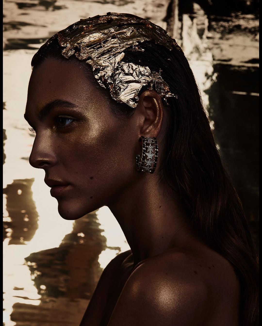 Portrait of woman with dark hair, gold leaf hair accessory, hoop earrings, and off-the-shoulder top against blurred gold and black background.