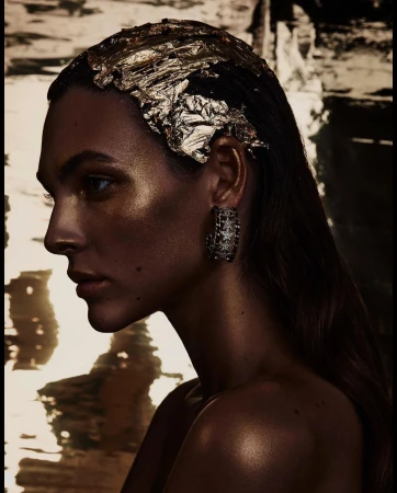 Portrait of woman with dark hair, gold leaf hair accessory, hoop earrings, and off-the-shoulder top against blurred gold and black background.