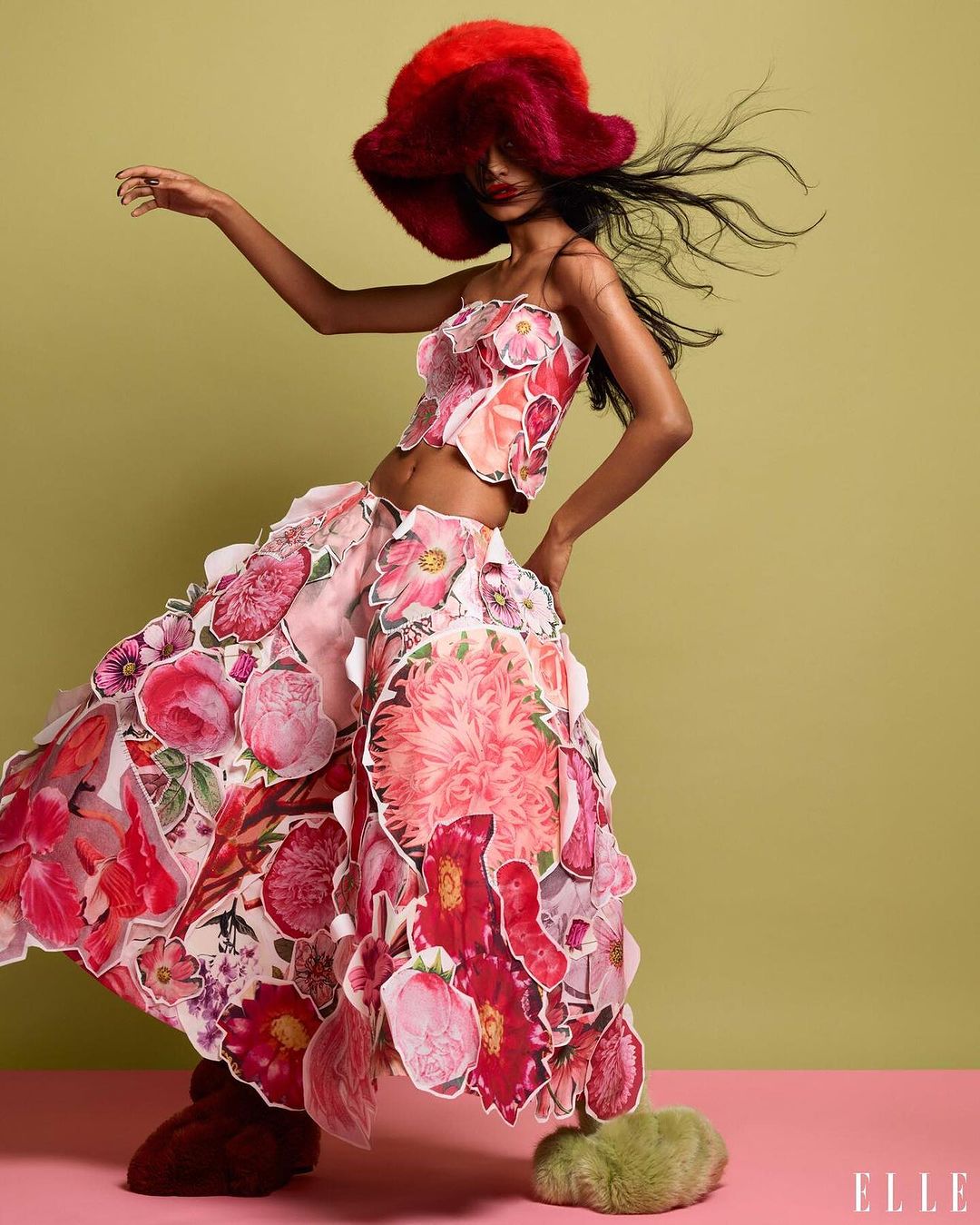 A runway fashion model strikes a dynamic pose wearing a floral-themed top and skirt with a large red hat against a green studio backdrop.