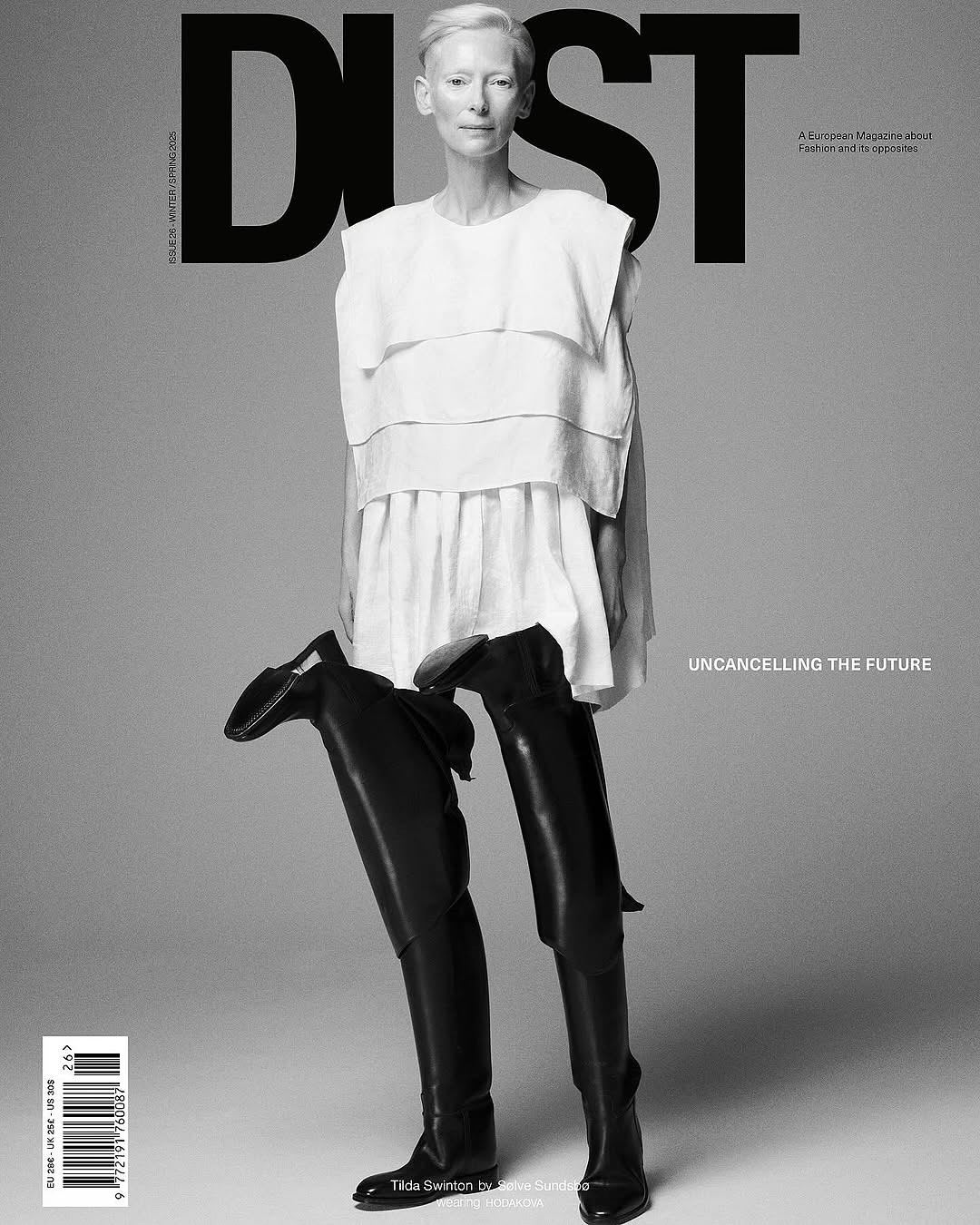 Black-and-white magazine cover featuring a fair-skinned woman in a white top and black thigh-high boots against a grey background.