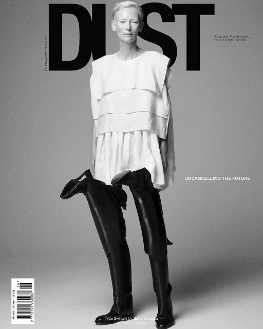 Black-and-white magazine cover featuring a fair-skinned woman in a white top and black thigh-high boots against a grey background.