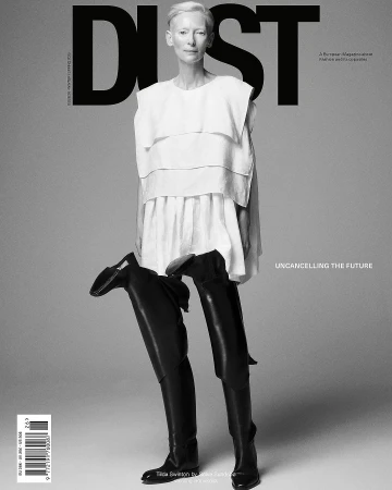Black-and-white magazine cover featuring a fair-skinned woman in a white top and black thigh-high boots against a grey background.