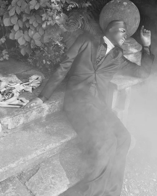 Black and white fashion photograph of a man in a suit lying on stone steps near leafy plants and a large stone sphere in the background.