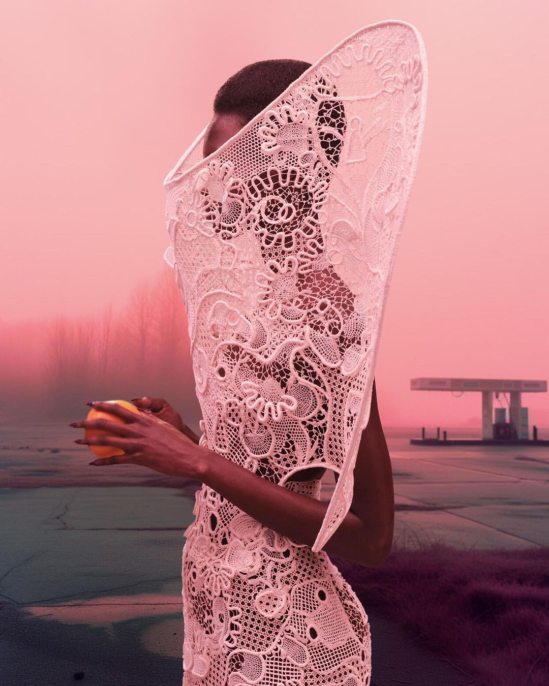 Woman in a white lace garment holds an orange in an outdoor setting with a pink sky and a distant gas station.