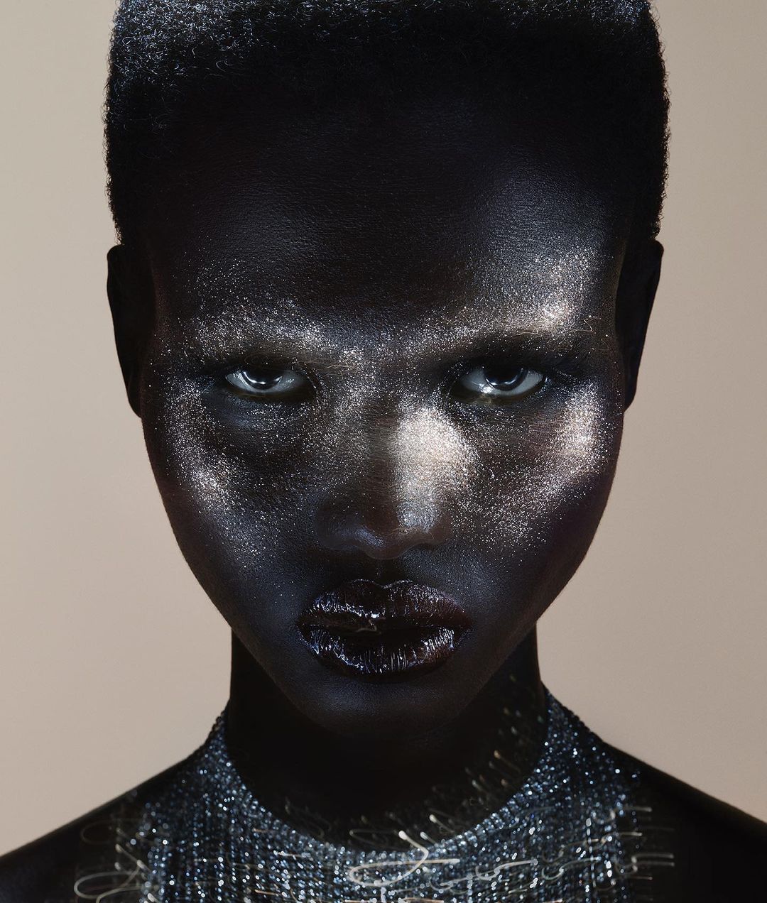 Close-up portrait of a woman with dark skin, silver-toned facial makeup, and a beaded necklace, set against a neutral, light-colored backdrop.