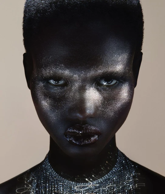 Close-up portrait of a woman with dark skin, silver-toned facial makeup, and a beaded necklace, set against a neutral, light-colored backdrop.