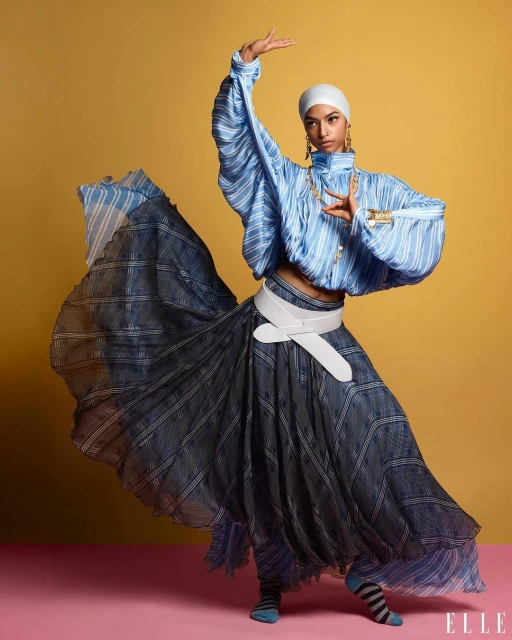 Elegant fashion shot: Woman in headscarf, flowing blue and black striped skirt, dances against a yellow and pink backdrop.