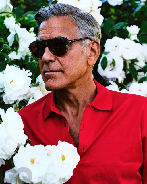 Portrait of George Clooney wearing sunglasses and a red polo shirt surrounded by white roses in an outdoor setting.