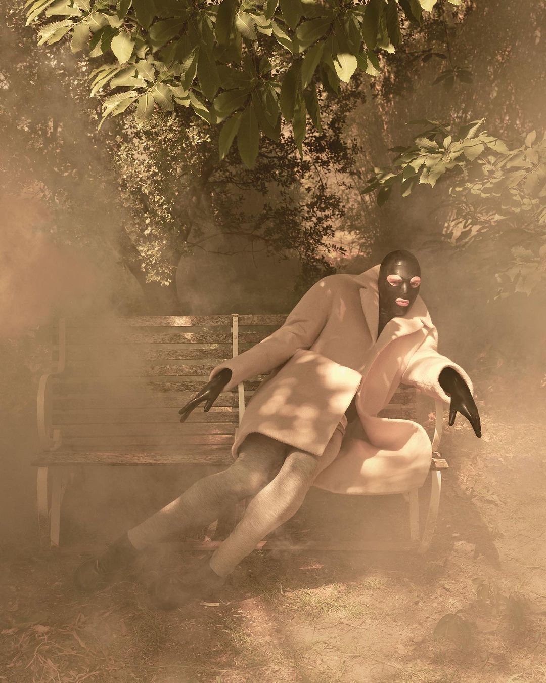 Figure in black latex mask and light coat sitting on park bench surrounded by atmospheric smoke by Sølve Sundsbø