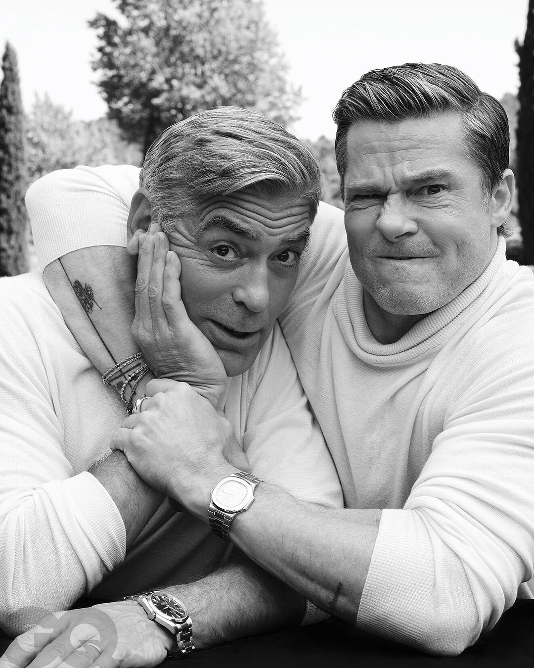 Black and white portrait of George Clooney and Brad Pitt posing together outdoors, both in turtlenecks and wearing wristwatches.