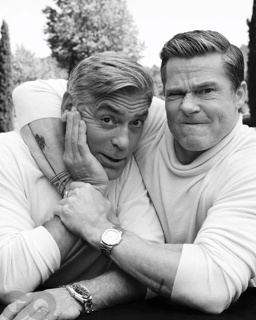 Black and white portrait of George Clooney and Brad Pitt posing together outdoors, both in turtlenecks and wearing wristwatches.