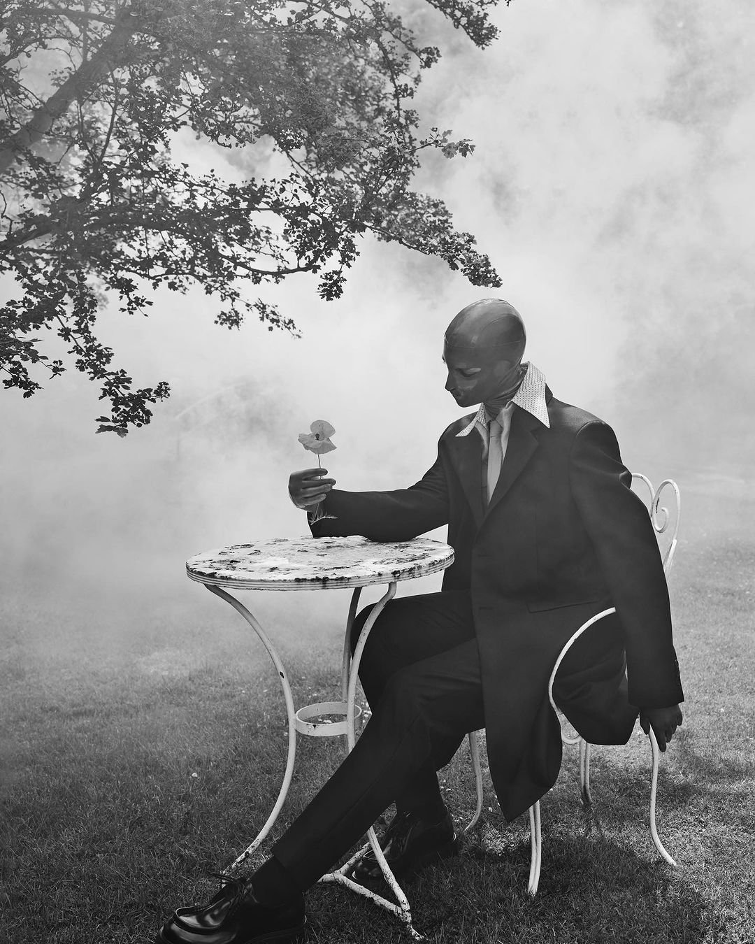Monochrome portrait of a seated man in a suit holding a flower outdoors with smoke or fog in the background by Solve Sundsbo