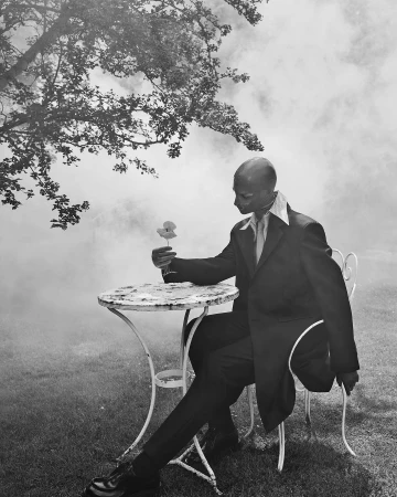 Monochrome portrait of a seated man in a suit holding a flower outdoors with smoke or fog in the background by Solve Sundsbo