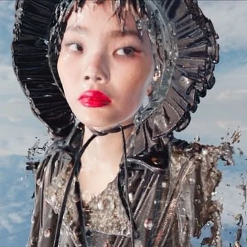 Dramatic headshot of an Asian woman with bold red lipstick wearing a shiny, futuristic hat and jacket against a cloudy blue sky backdrop.
