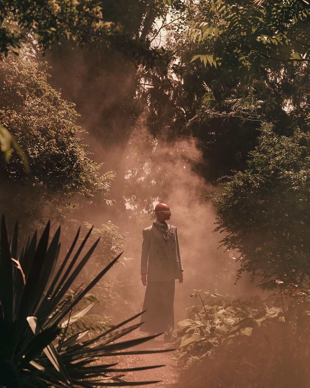 An adult with shaved head in a formal grey suit and long skirt stands in a lush forest shrouded in mist, creating a surreal and captivating image.